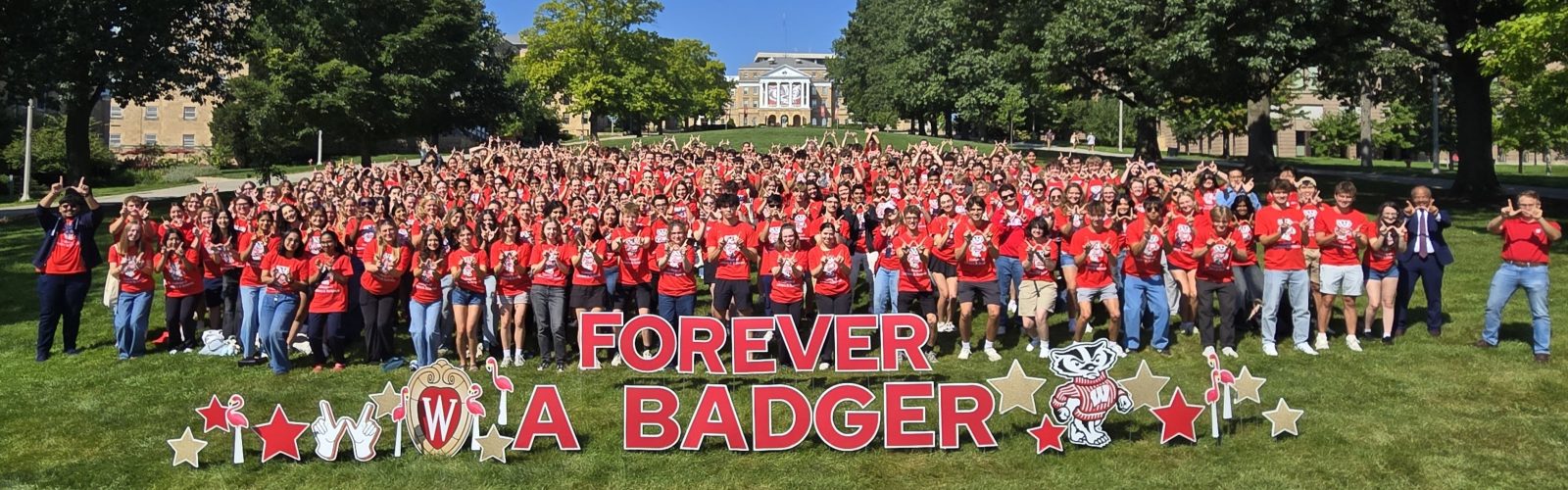 Over 300 incoming First Year L&S Honors Students in red shirts gather on Bascom Hill behind "Forever A Badger" signage.