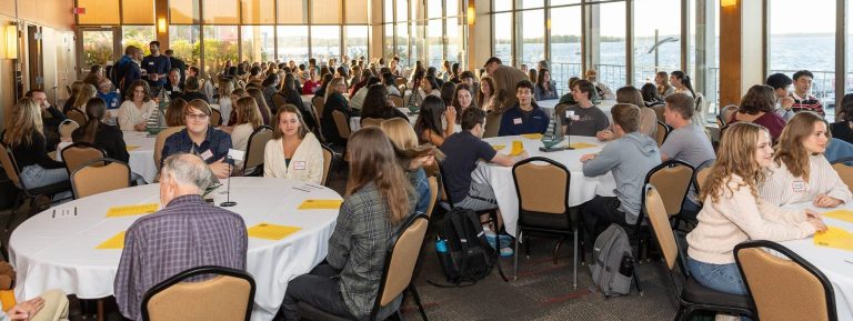 Large room overlooking a lake with many round tables. Students converse with faculty.