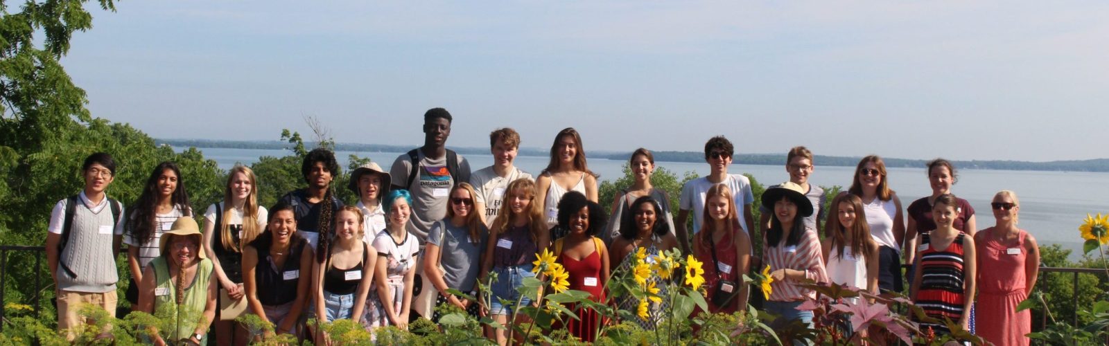 Group of 26 students with nametags smiling in front of plants and a lake.