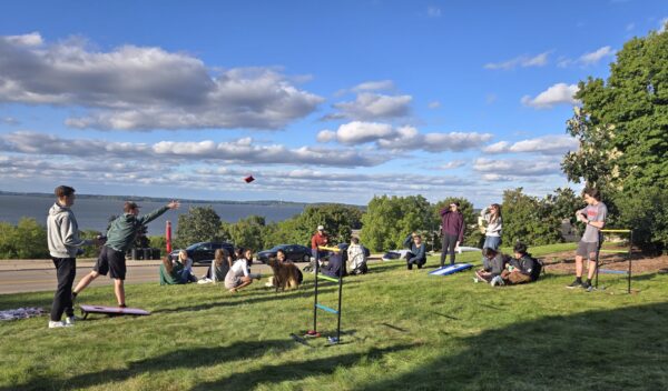 Students throw beanbags in front of other students sitting on the grass, a dog, and a background of lake, sky, and trees.