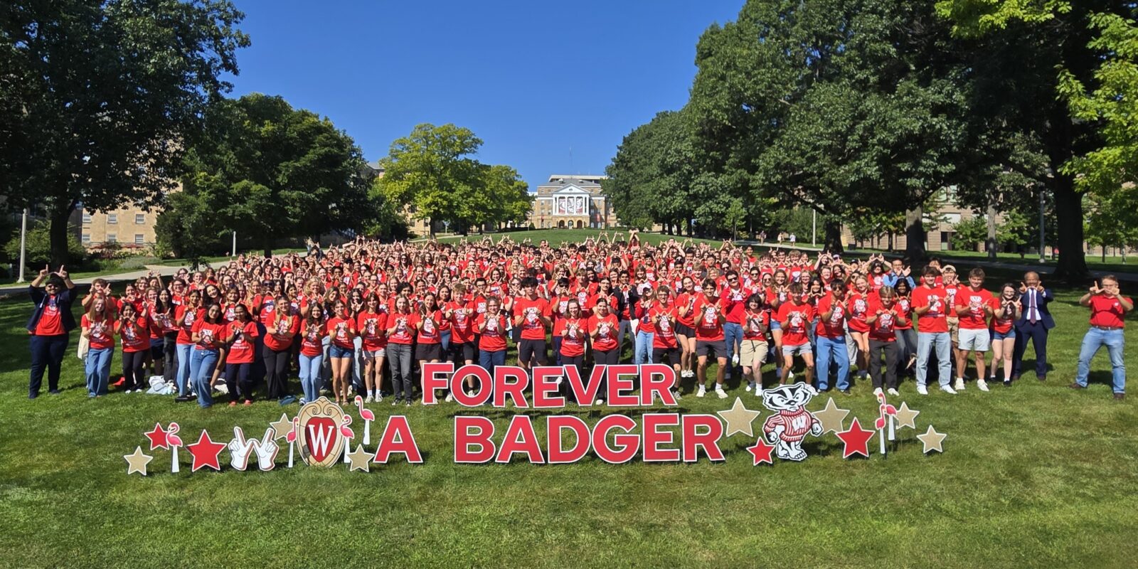 Many students in red shirts smile for picture on a grassy hill with a "Forever a Badger" sign in the foreground
