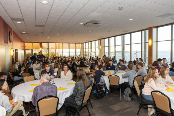 Large room with many people seated at round tables. Large windows on right side look out over a lake.